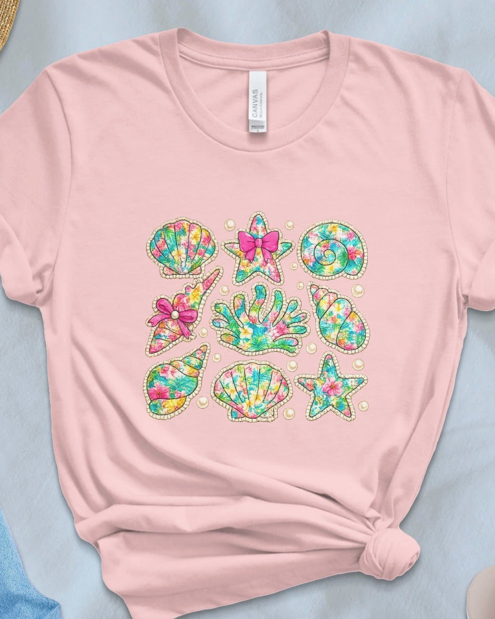 Pink t-shirt with colorful mermaid-themed designs on a light blue surface with accessories.