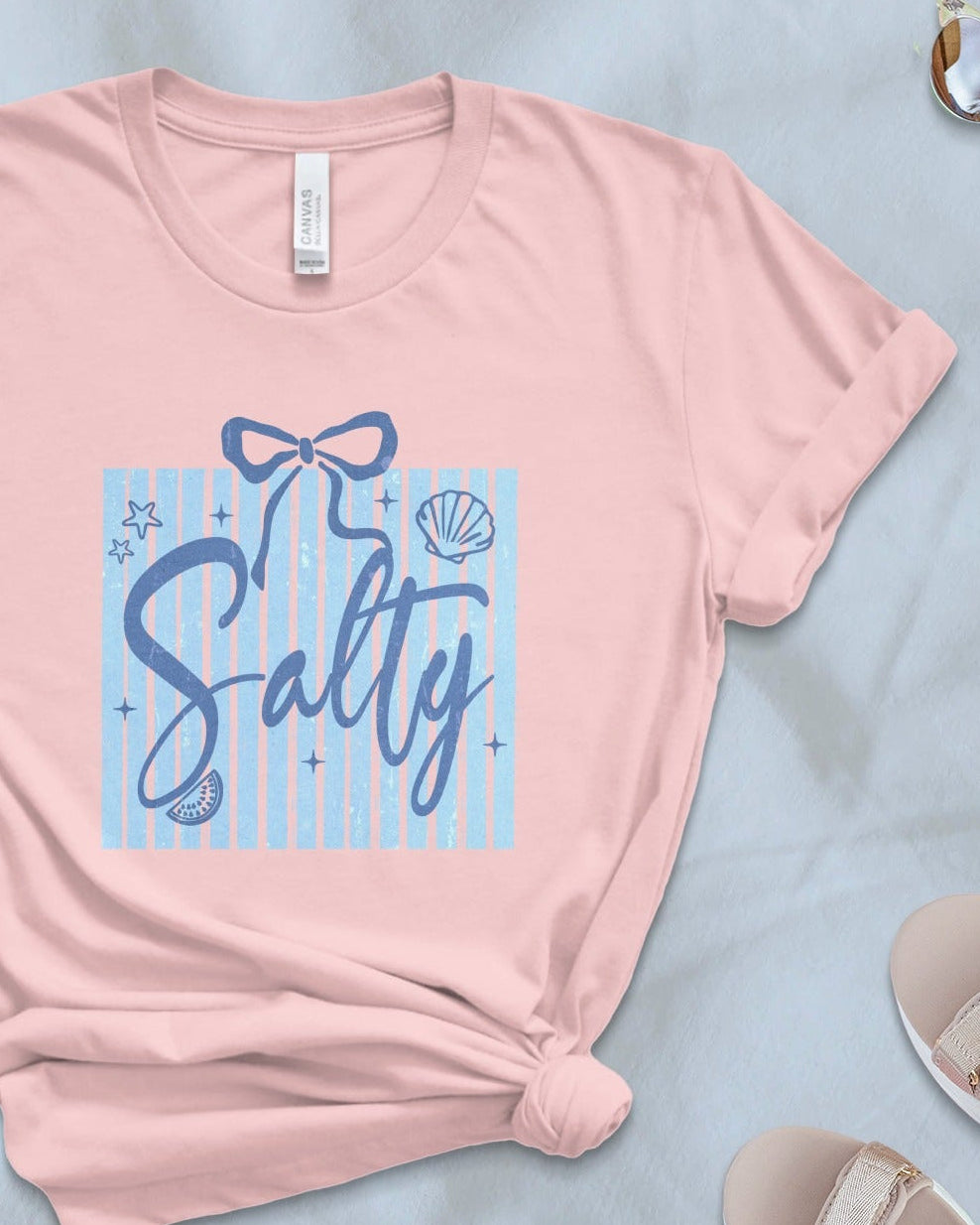Pink t-shirt with 'Salty' design on a light blue surface with accessories.