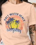 Person wearing a peach t-shirt with a palm tree and sunset design and text 'Salt Water Heals Everything'.