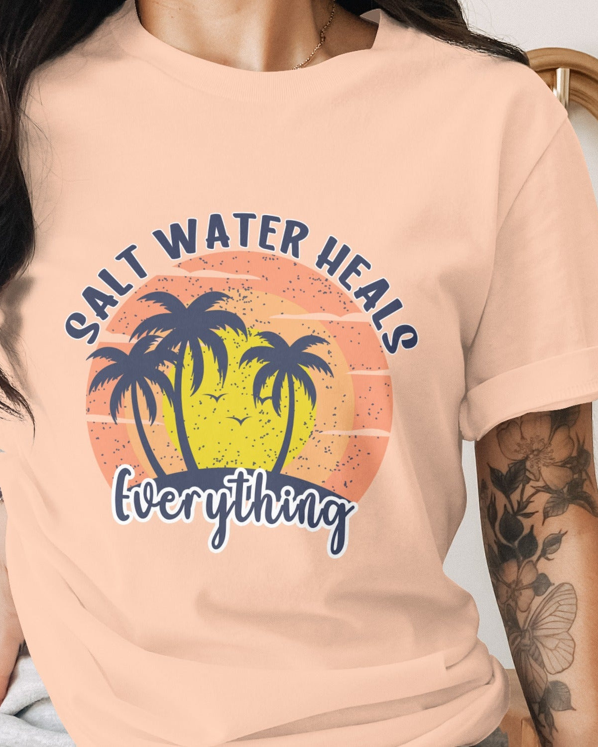 Person wearing a peach t-shirt with a palm tree and sunset design and text 'Salt Water Heals Everything'.