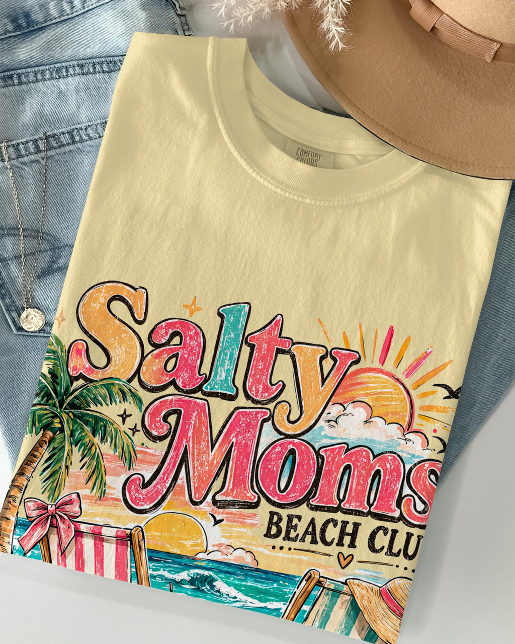 Beige t-shirt with 'Salty Moms Beach Club' design, denim jacket, and beige hat on a light gray background