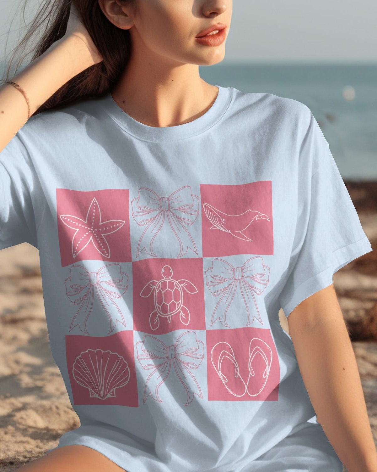 Woman wearing a light blue t-shirt with pink and white graphic design on a beach.