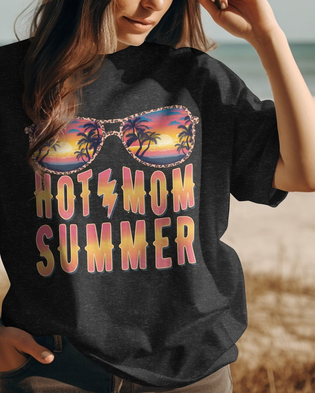 Person wearing a black t-shirt with 'HOT MOM SUMMER' and sunglasses graphic on a beach.