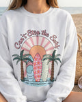 White sweatshirt with a graphic design of palm trees, surfboards, and text 'Can't Stop the Surf' worn by a person.