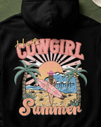Black hoodie with a 'Hot Cowgirl Summer' graphic design featuring a woman with a surfboard on a beach.