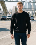 A man walking on a sunny city sidewalk in front of a large arched bridge, wearing a black crewneck sweatshirt, dark blue jeans, and white sneakers.