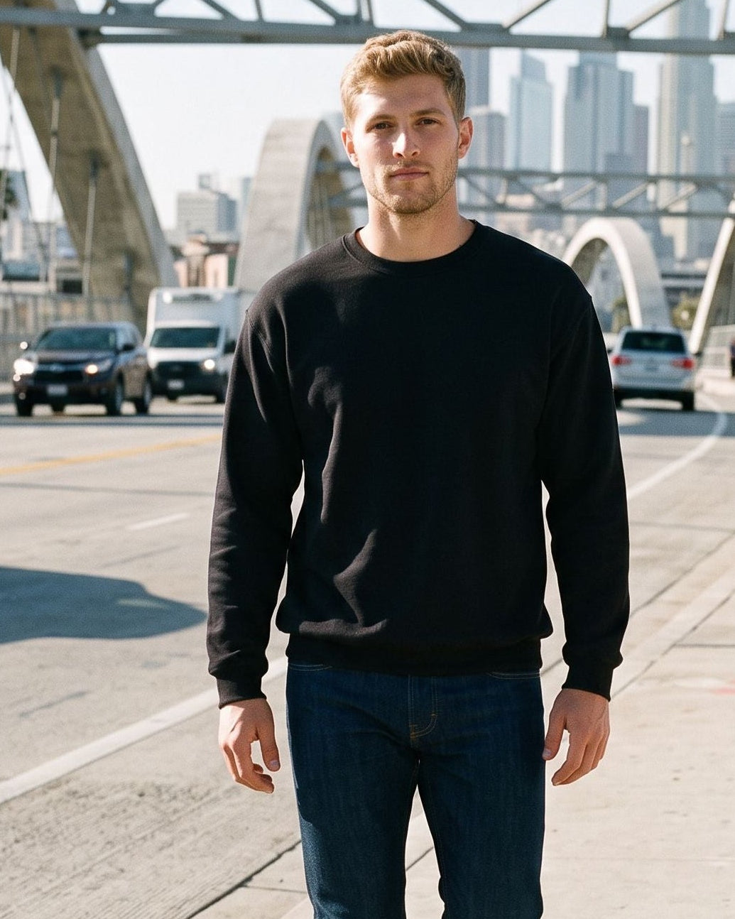 A man walking on a sunny city sidewalk in front of a large arched bridge, wearing a black crewneck sweatshirt, dark blue jeans, and white sneakers.