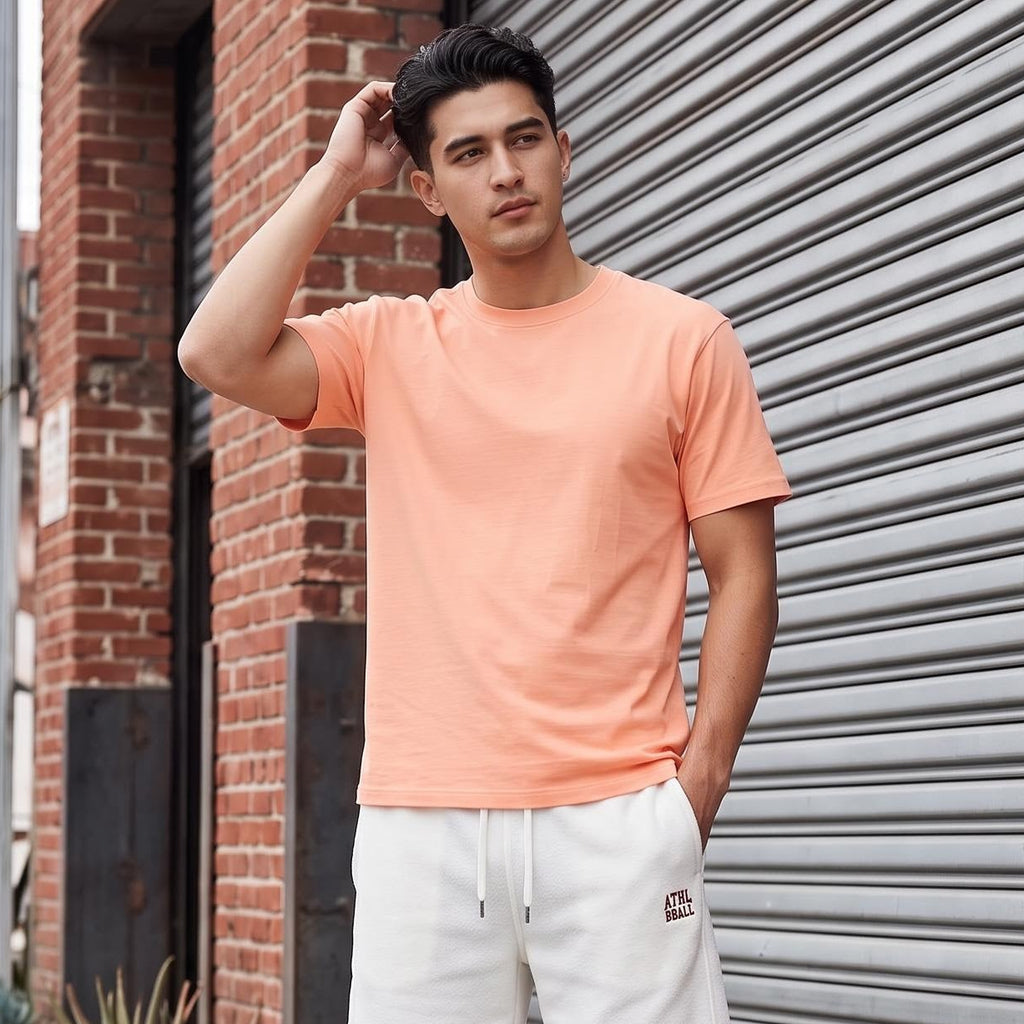 A man stands on a city sidewalk wearing a peach crewneck t-shirt, off-white drawstring shorts with a small red circular patch, and white sneakers, positioned in front of a gray metal roll-up garage door and brick wall.