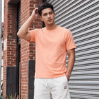 A man stands on a city sidewalk wearing a peach crewneck t-shirt, off-white drawstring shorts with a small red circular patch, and white sneakers, positioned in front of a gray metal roll-up garage door and brick wall.