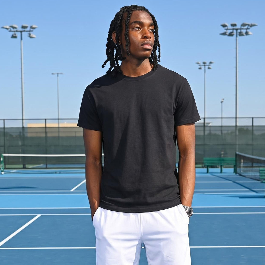 A man stands on a blue tennis court wearing a black short-sleeve t-shirt, white athletic shorts, white socks, and white sneakers.