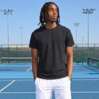 A man stands on a blue tennis court wearing a black short-sleeve t-shirt, white athletic shorts, white socks, and white sneakers.