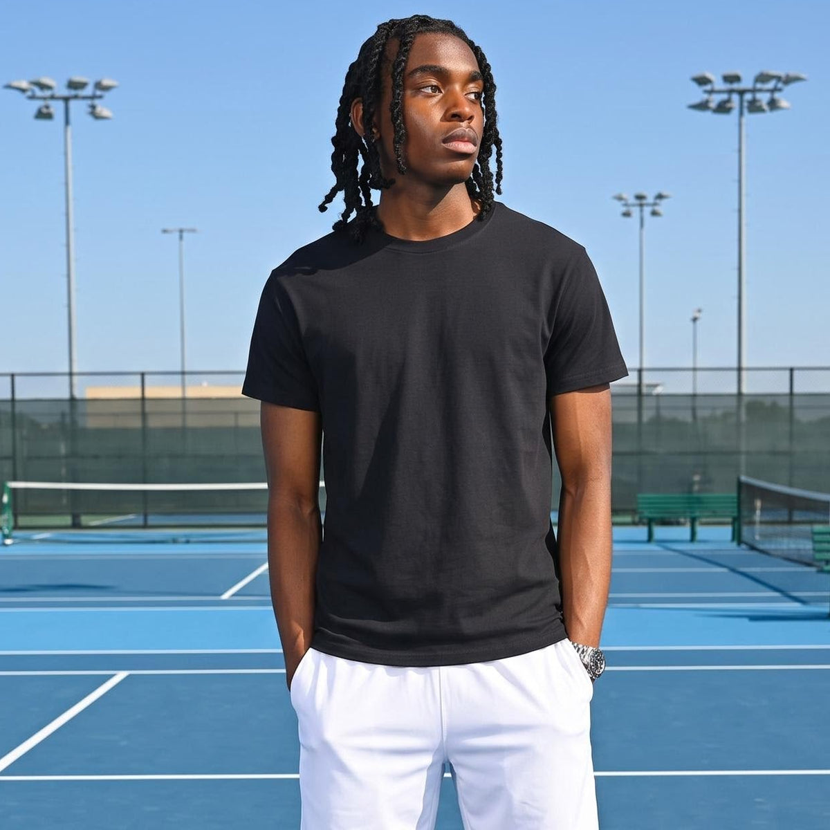 A man stands on a blue tennis court wearing a black short-sleeve t-shirt, white athletic shorts, white socks, and white sneakers.